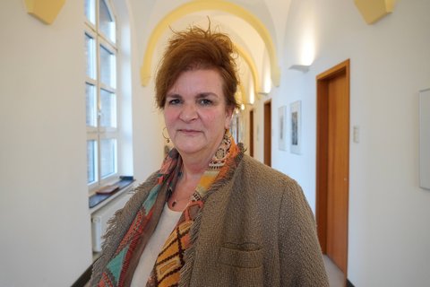 Eine Frau mit hochgesteckten Haaren und einem leichten Schal um den Hals steht auf einem beleuchteten Flur.