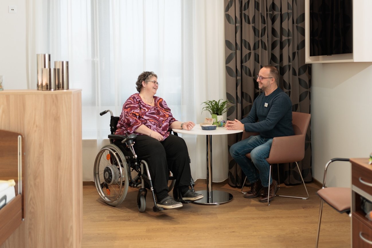 Ein Fachkrankenpfleger sitzt in einem Patientenzimmer und berät an einem runden Tisch lächelnd eine Frau im Rollstuhl im Rahmen seiner Aufgaben der Psychiatrische Pflege Köln. 