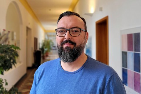 Ein Mann mit Kinnbart, schwarzer Brille und blauem T-Shirt steht in einem sanf beleuchteten Flur. Man sieht Pflanzen und Bilder im Hintergrund. 