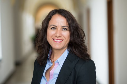 Eine Frau mit schwarzen langen Haaren und schwarzer Jacke steht auf einem beleuchteten Flur